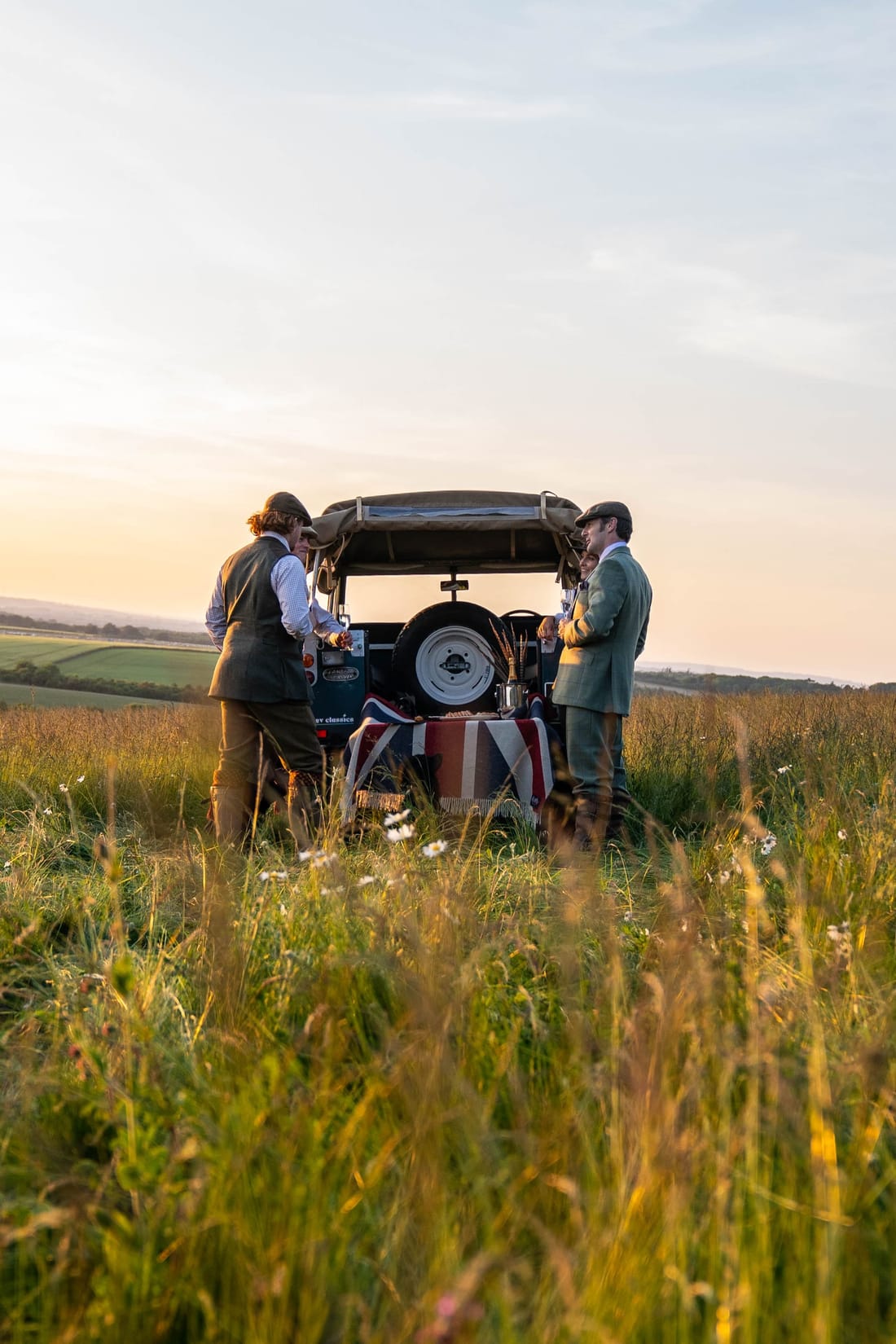 Two men in country tweeds with the EV Classics Series 3 at sunset, Union Jack picnic blanket draped from the tailgate, on the Dege & Skinner shoot